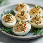 Deviled eggs filled with a textured everything bagel mixture and topped with chives on a gray plate with herbs.