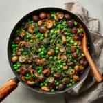 Top-down skillet filled with peas, golden mushrooms, herbs, and a wooden spoon on a linen cloth.