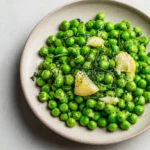 Top-down view of bright green peas mixed with lemon wedges and herbs on a ceramic plate.