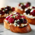 Crisp crostini topped with creamy goat cheese, roasted red grapes, and fresh herbs on a bright marble surface.