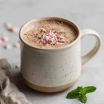 A ceramic mug of peppermint hot cocoa sprinkled with crushed candy canes on top, styled on a light surface with a fresh mint sprig nearby.