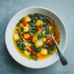 Top view of a vegetable soup bowl with carrots, potatoes, kale, and herbs on a cool-toned background.