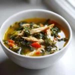 White bowl of chicken and kale soup on a bright surface, filled with leafy greens, carrots, and tender chicken.