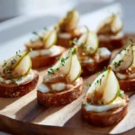 Caramelized Pear & Brie Canapés 3 Crostini topped with fresh pear slices, melted brie, thyme leaves, and a light drizzle of caramel sauce on a wooden board.