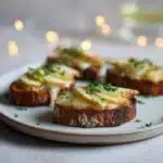 Smoked Gouda & Apple Crostini with Honey Drizzle 3 Toasted crostini with melted gouda and thin apple slices sprinkled with fresh herbs on a ceramic plate.