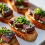 Toasted crostini topped with thick seared steak slices and fresh herbs arranged on a bright white plate.