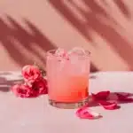 Valentine’s Love Potion Mocktail 3 Pink iced mocktail in a short glass surrounded by rose petals on a pink surface with soft shadows.