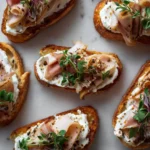 Smoked Turkey & Whipped Goat Cheese Toasts 3 Overhead view of multiple smoked turkey and whipped goat cheese toasts arranged on a light surface with herbs