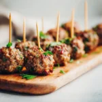 Baked mini meatball skewers lined on a wooden board with toothpicks, garnished with chopped herbs in natural light.