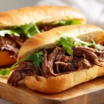 Shredded braised beef sandwiches on soft rolls with fresh herbs, arranged on a wooden board in bright natural light.