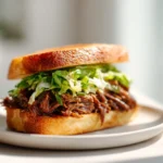 Beer-Braised Beef Sandwiches (Non-Alcoholic) 3 Close-up of a braised beef sandwich on toasted bread with shredded beef and fresh greens on a white plate.