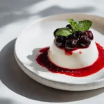 Single vanilla panna cotta on a white plate with berry coulis pooled around the base and fresh berries on top.
