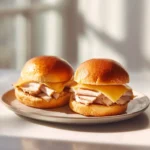 Ham and Cheese Hawaiian Sliders 3 Two Hawaiian roll sliders with sliced turkey and cheese on a ceramic plate near a sunlit window.