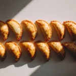 Row of baked chicken empanadas arranged neatly on a light surface with warm natural light.