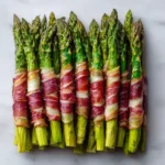 Overhead view of neatly lined asparagus spears wrapped in beef pancetta, roasted golden and arranged on a light surface.