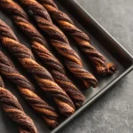 baked puff pastry chocolate twists lined up on a dark baking tray with deep chocolate color and crisp twisted texture