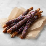 stacked chocolate dipped pretzel rods with rainbow sprinkles resting on parchment paper