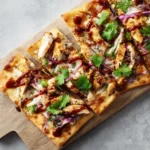 Rectangular BBQ chicken flatbread sliced into squares with red onion, cilantro, and BBQ drizzle on wooden board.