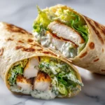 Close-up of sliced chicken Caesar wrap with grilled chicken, romaine, and creamy dressing on marble surface.