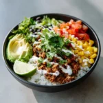 Ground beef burrito bowl with rice, avocado slices, corn, black beans, pico de gallo, cilantro, and sour cream drizzle.