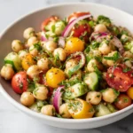 Spring Chickpea Salad with Fresh Herbs 3 Close-up of chickpea salad with yellow and red tomatoes, cucumber, red onion, and chopped herbs in a white bowl.