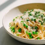 Bowl of spaghetti with peas, Parmesan cheese, and fresh thyme on a light kitchen surface.