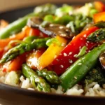 Close-up of glossy spring vegetable stir-fry with asparagus, bell peppers, broccoli, and mushrooms over brown rice in a dark bowl.