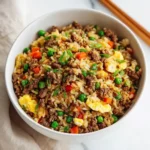 Bowl of ground beef fried rice topped with scrambled eggs and green onions on a light surface.