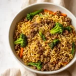 White bowl of ground beef ramen noodles with broccoli and carrots on a light surface.