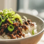 Ground beef teriyaki served over white rice in a ceramic bowl, topped with sliced green onions and sesame seeds in natural light.