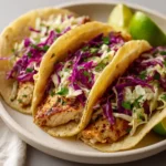 Three chicken tacos topped with purple and green cabbage slaw on a neutral plate with lime wedges.