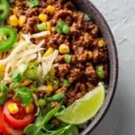 Ground beef burrito bowl with rice, black beans, corn, jalapeños, shredded cheese, tomatoes, and lime wedge.