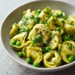 Bowl of pesto tortellini with peas topped with cracked black pepper and fresh herbs on a neutral background