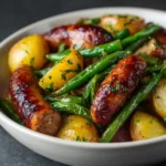 One-Pan Sausage with Potatoes and Green Beans 3 Roasted sausage links with baby potatoes and green beans served in a white bowl.