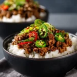 Teriyaki ground beef over rice topped with sliced jalapeños, red chili peppers, and sesame seeds in a dark bowl.