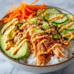 Teriyaki-style salmon rice bowl with avocado, cucumber, shredded carrots, sesame seeds, and creamy sauce drizzle.